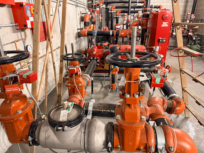 Mechanical room with a network of orange and black pipes, valves with gauges, and fire safety equipment.