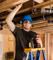 Man wearing blue safety helmet and glasses standing on ladder while working on ceiling pipes in a basement.
