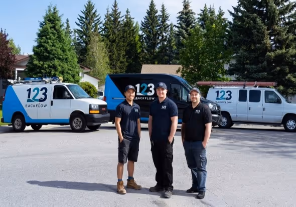 Three men in black company shirts standing in a parking lot with three white and blue vans labeled '123 BACKFLOW' in the background.