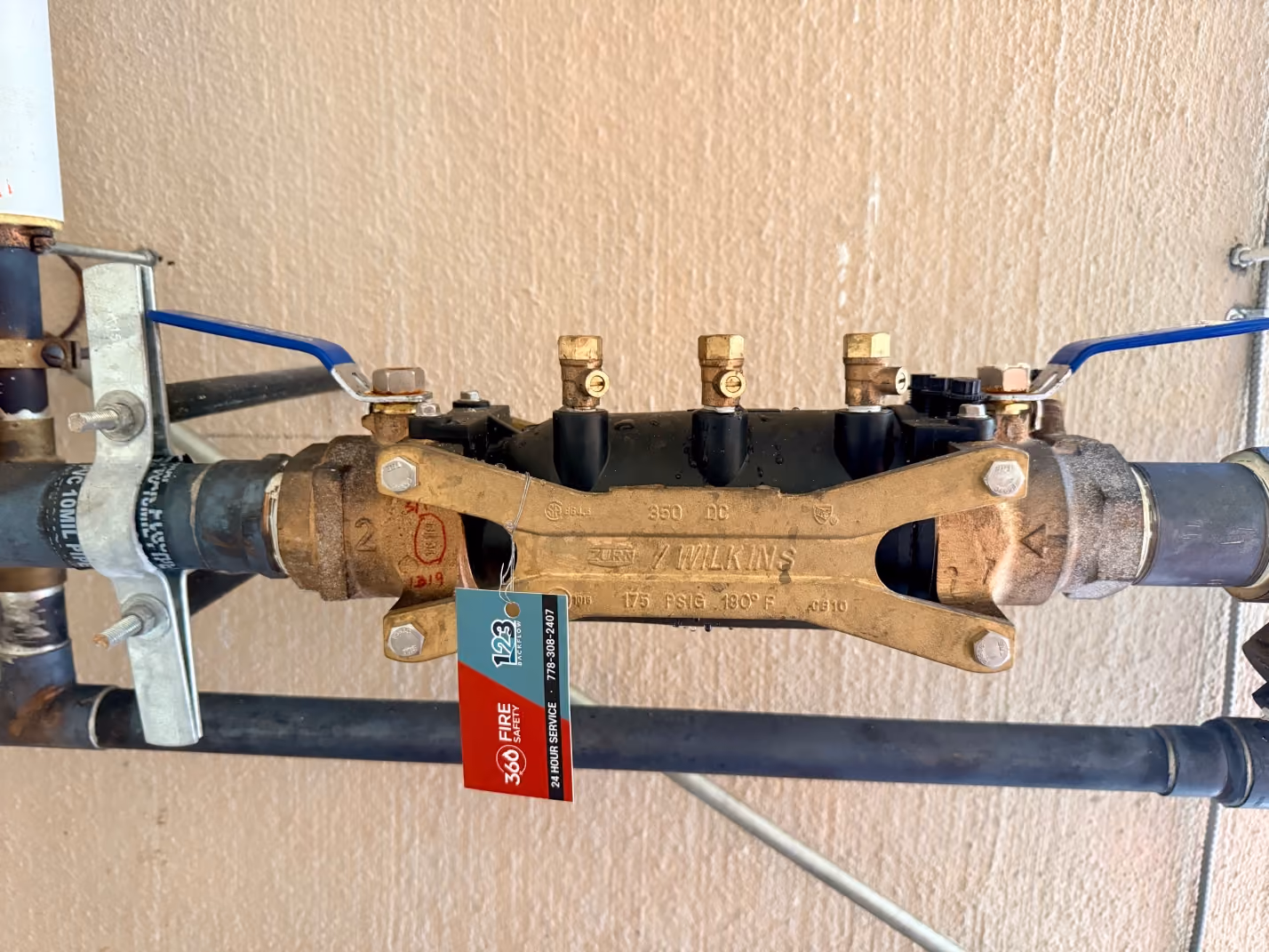 Brass fire safety valve with two blue handles and attached 360 Fire Safety tag on wall pipes.