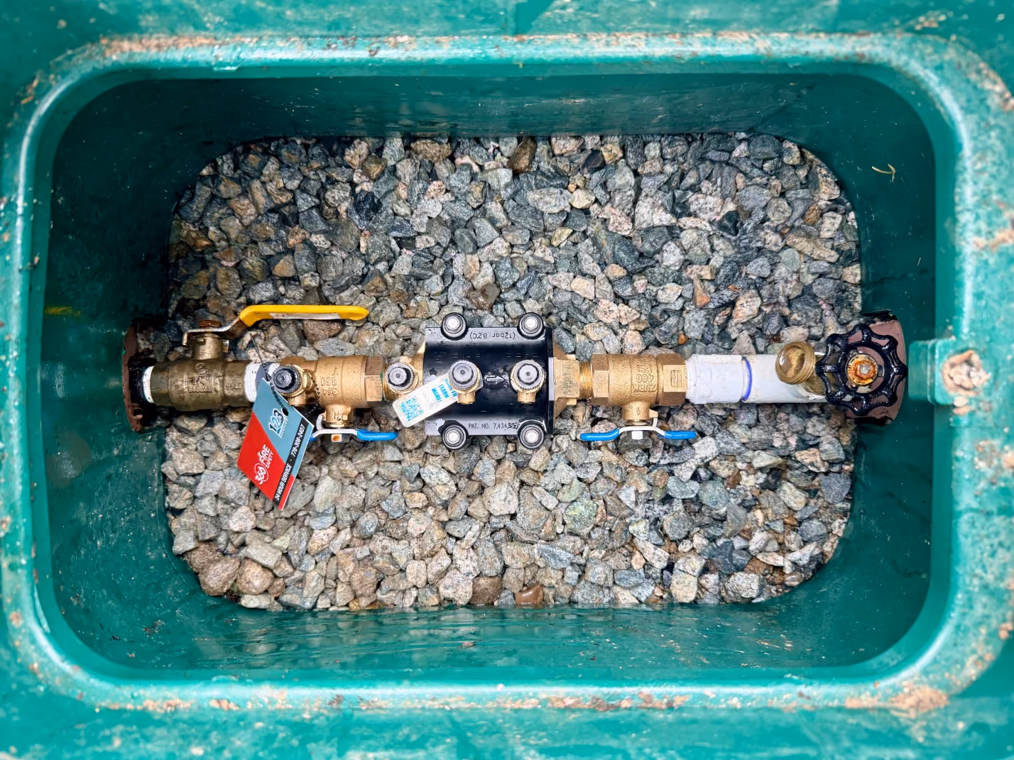 Water meter assembly with valves set in a green box filled with gravel.