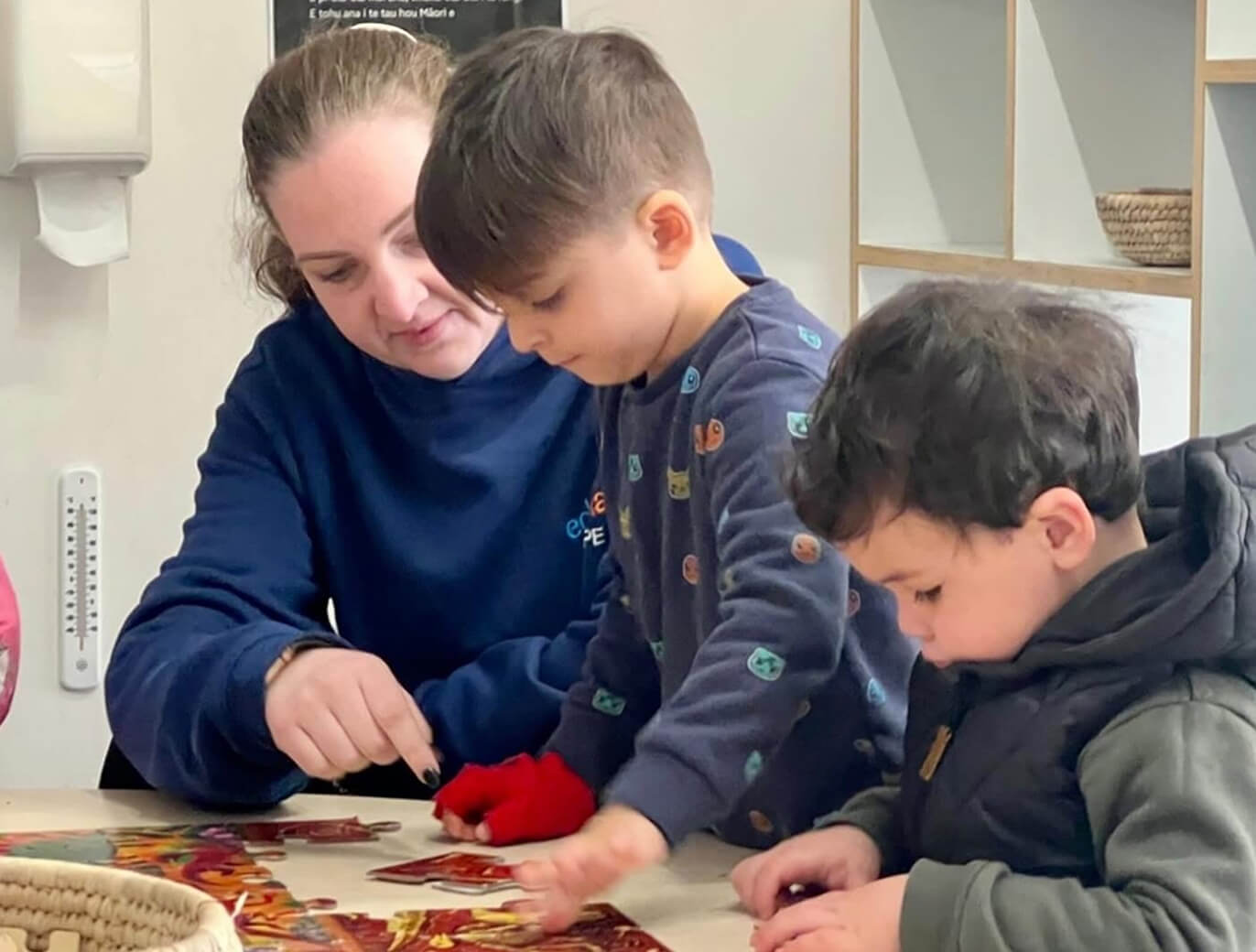 Adult woman and two young boys assembling a colorful jigsaw puzzle on a table indoors.