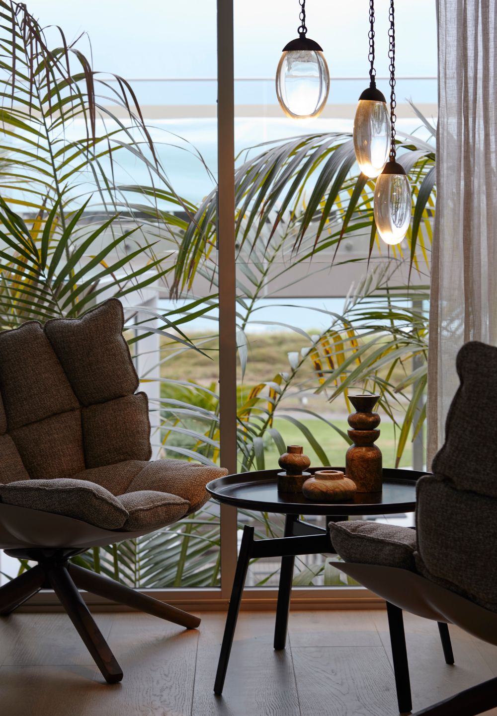 Omaha Retreat living area with upholstered lounge chairs, timber flooring and pendant lighting framed by the coastal landscape.