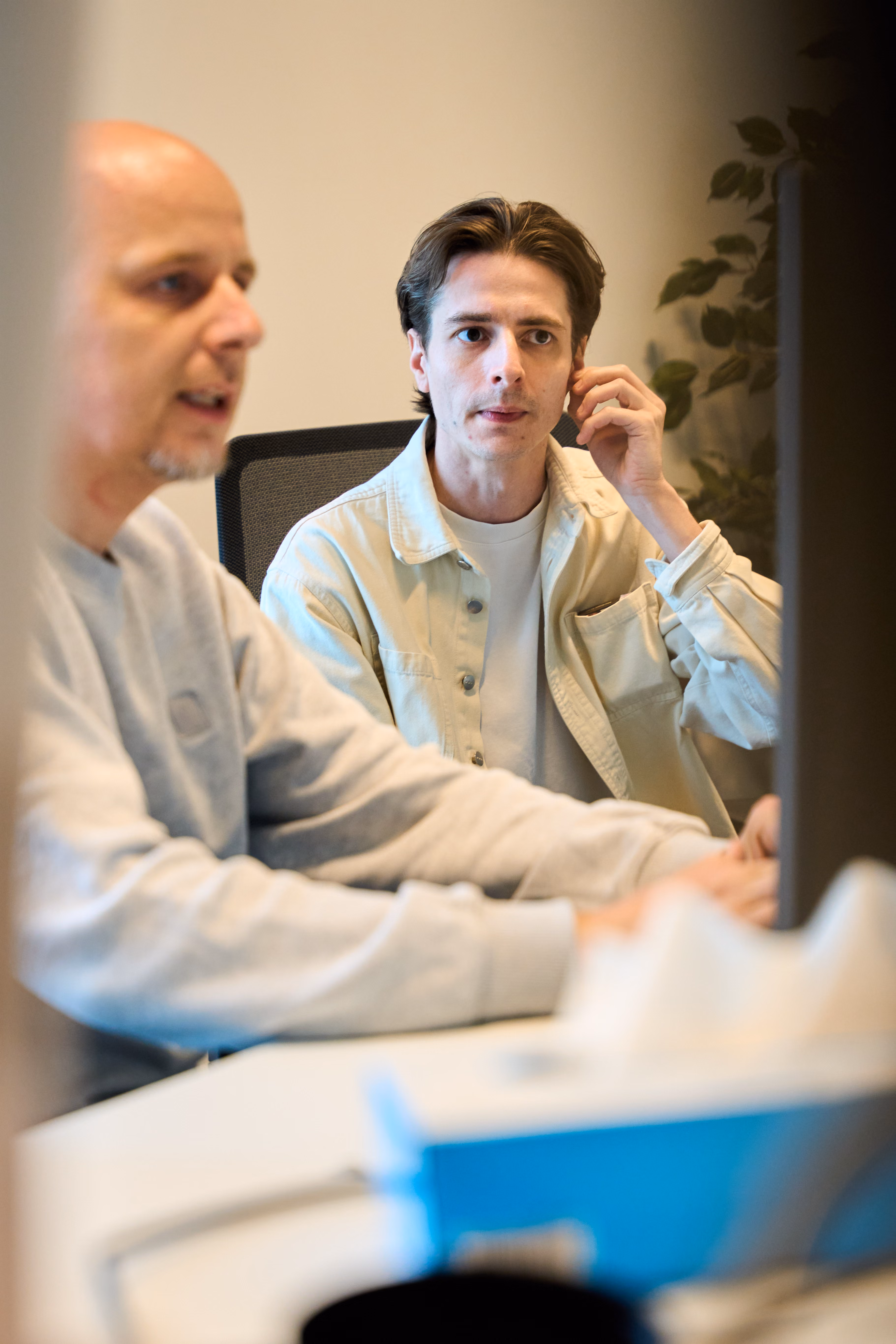 Two colleagues seated at a meeting table, one man in a blue shirt smiling and gesturing, and one woman with tattoos listening attentively.