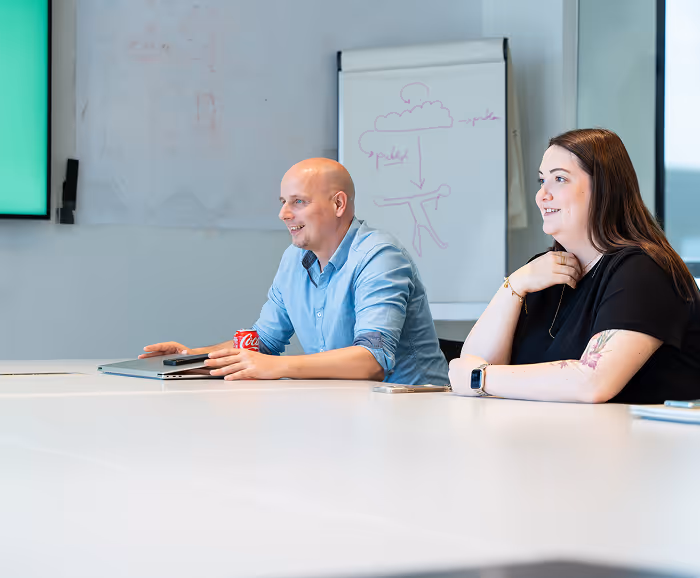 Two colleagues seated at a meeting table, one man in a blue shirt smiling and gesturing, and one woman with tattoos listening attentively.