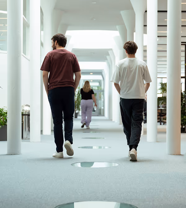 Three people walking down a bright office corridor with white pillars and large windows, two men in front and one woman in the back.