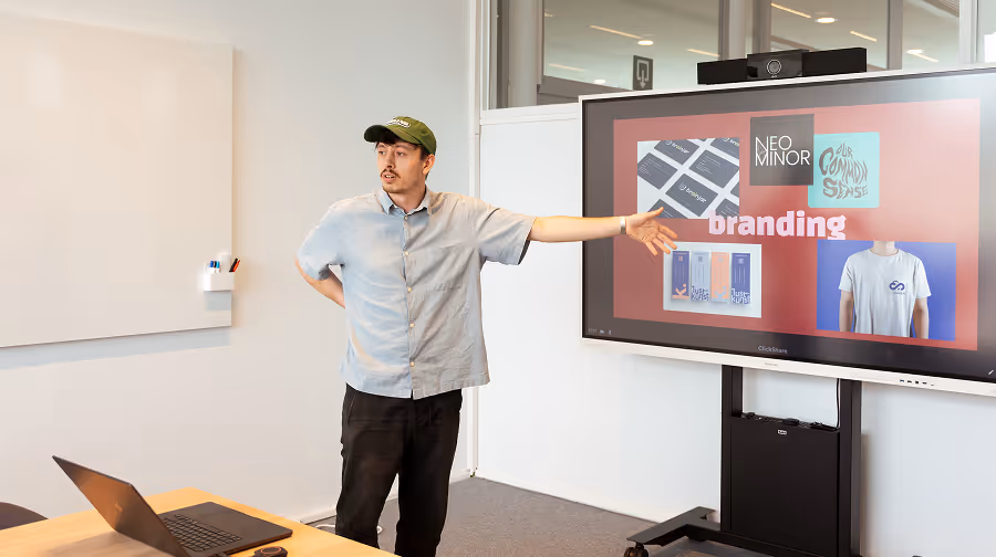 Man wearing a cap and light shirt presenting branding designs displayed on a large screen in an office meeting room.