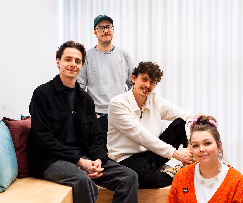 A group of four young adults smiling indoors against a white curtain background; three men and one woman seated on wooden benches with colorful cushions.
