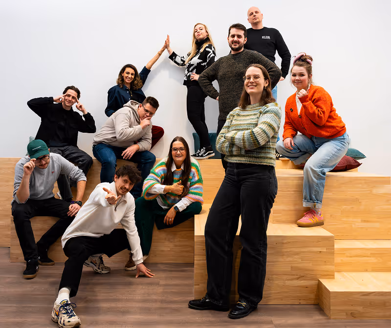 Group of eleven young adults posing happily on wooden steps against a white wall, with varied casual clothing and cheerful expressions.