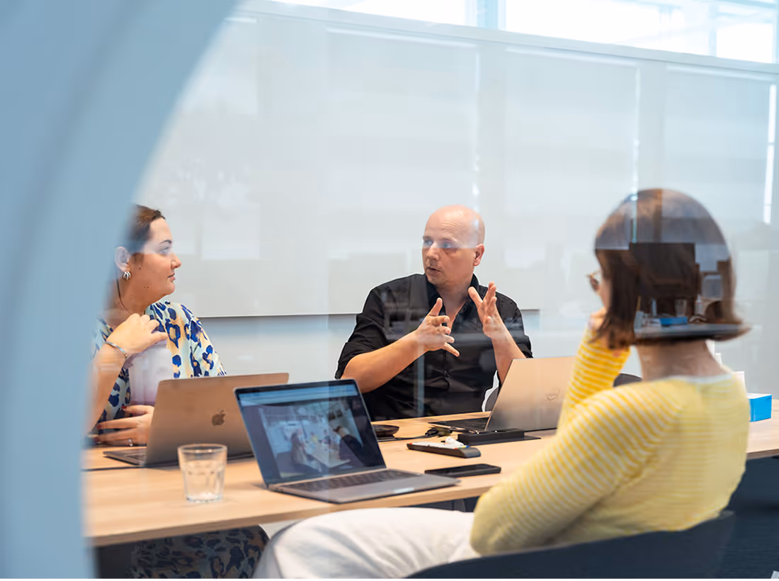 Three people having a discussion around a table in a modern office meeting room with laptops open.