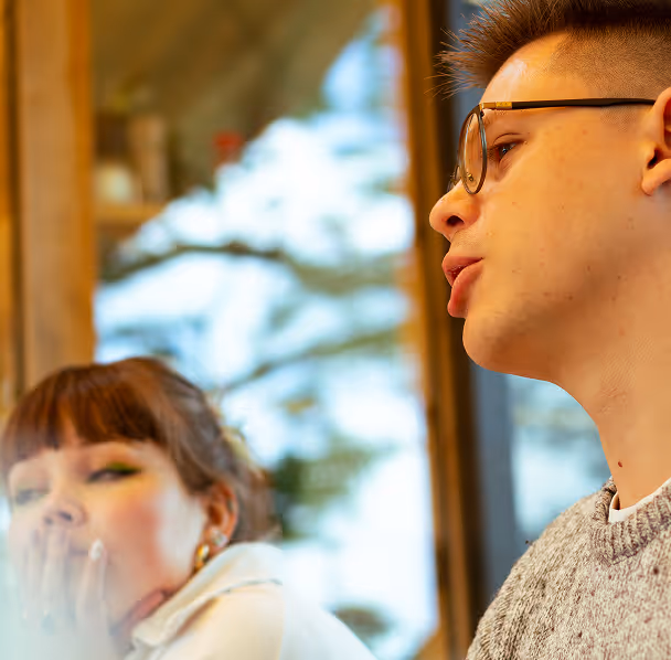Close-up of a young man wearing glasses speaking, with a blurred woman in the background covering her mouth.