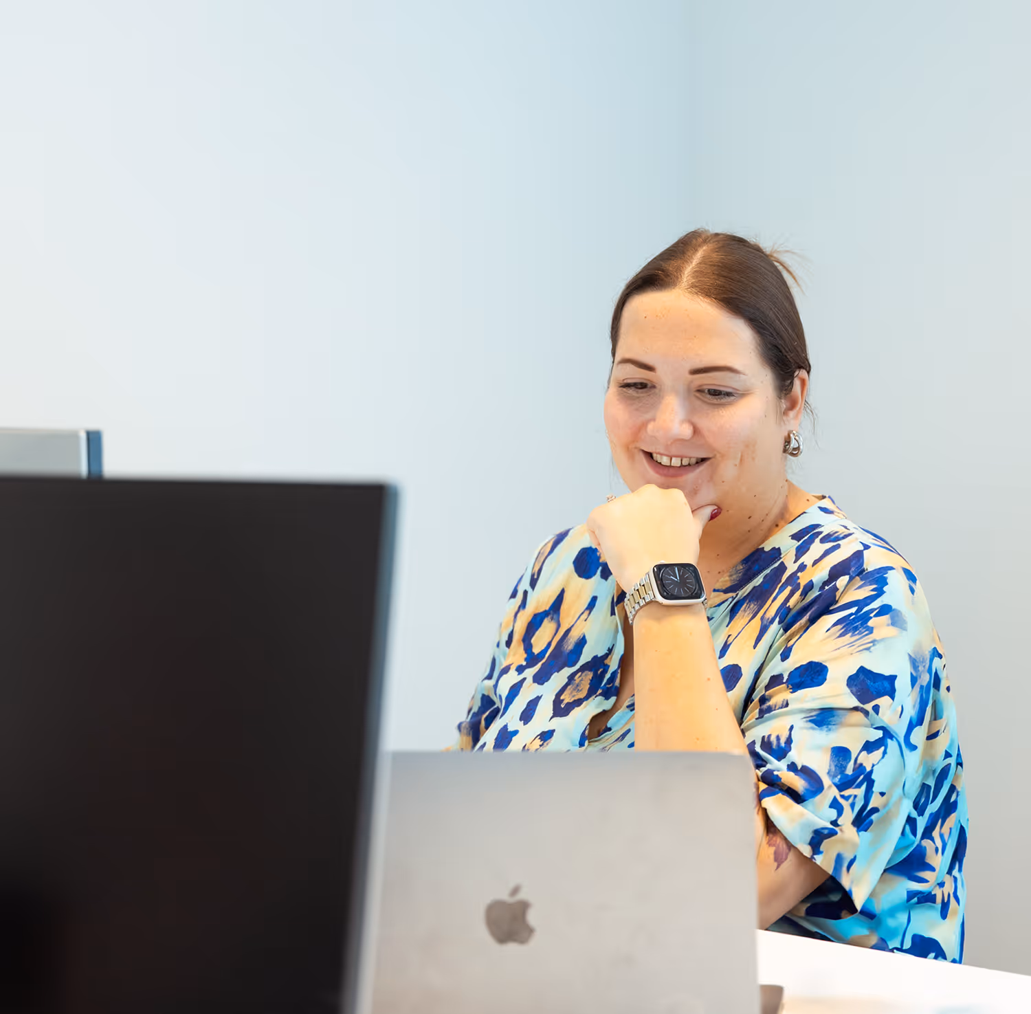 Smiling woman in a blue patterned blouse working on a laptop with an Apple logo.