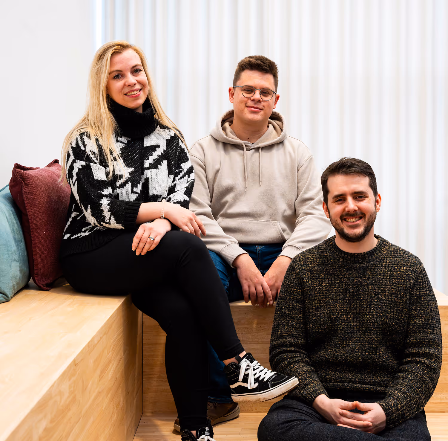 Three young adults smiling and sitting on wooden benches with colorful pillows behind them.