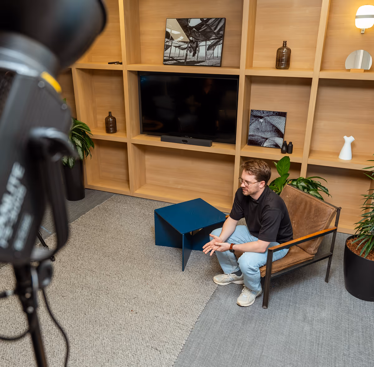 Man wearing glasses and casual clothes sitting on a brown chair in a modern living room with wooden shelves, a blue coffee table, and a large TV.