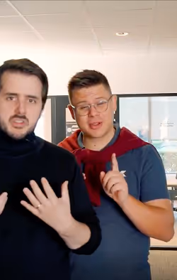 Two men indoors, one in a black top speaking with hands raised, the other in glasses and a blue shirt with a red sweater over shoulders gesturing with his finger.