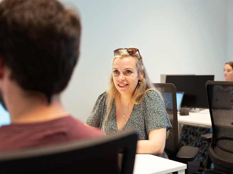 A woman with sunglasses on her head smiling and talking to a man in an office setting with computers in the background.