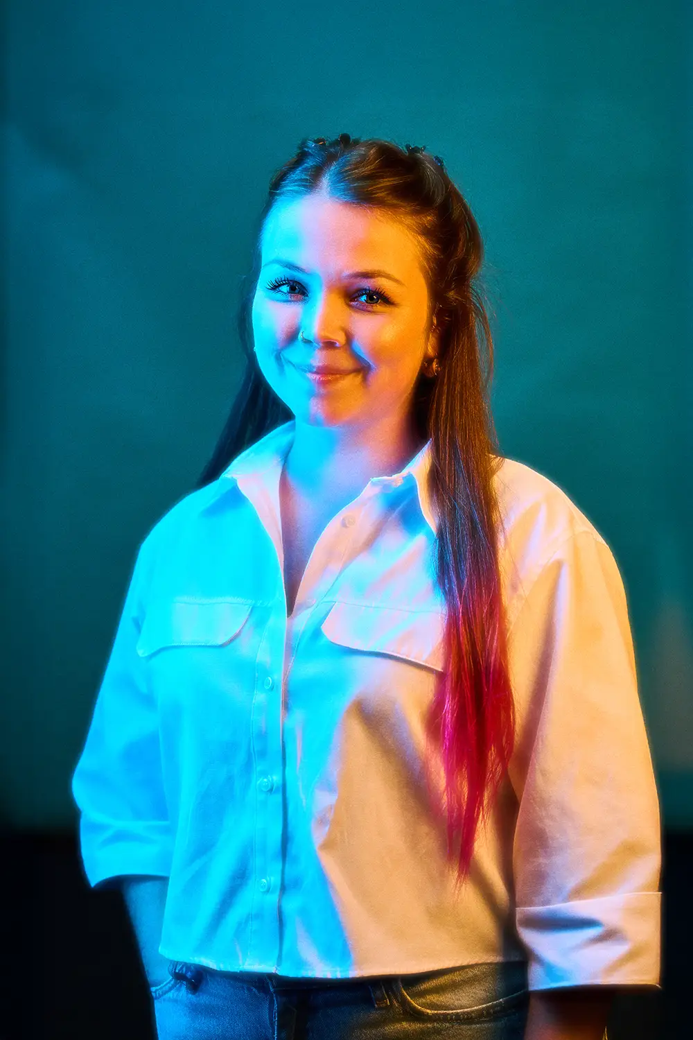 Smiling woman with long hair dyed pink at the ends, wearing a white button-up shirt against a teal background.