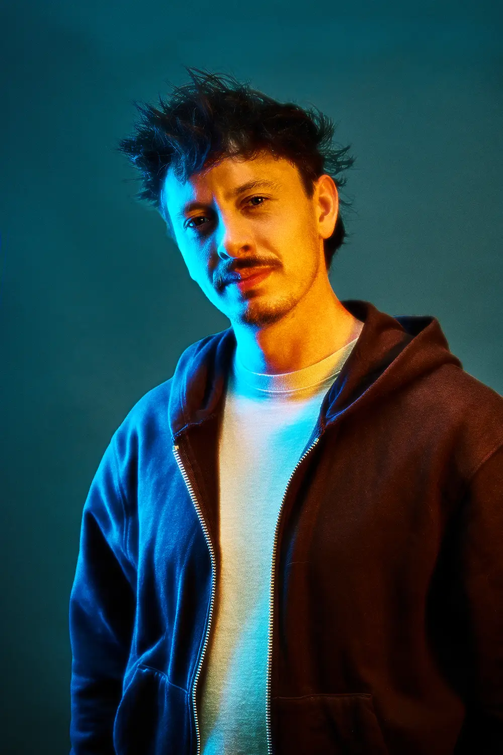 Young man with tousled hair wearing a white shirt and dark zip-up hoodie illuminated by blue and orange lighting against a dark background.