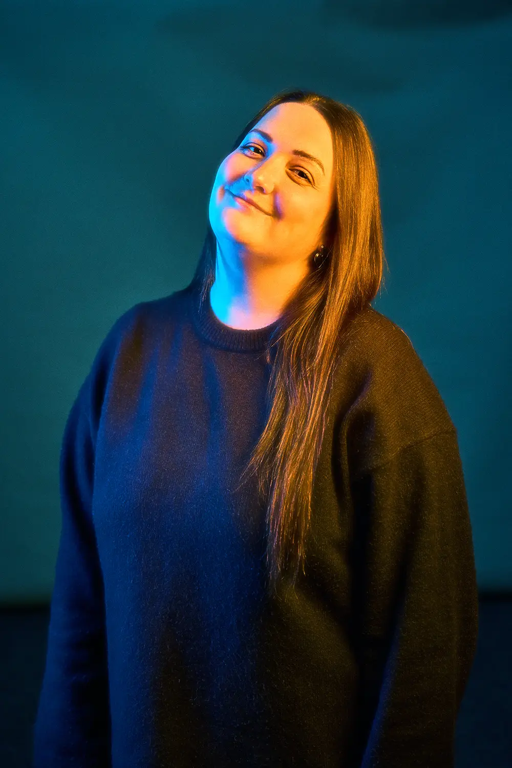 Smiling woman with long hair wearing a dark sweater illuminated by blue and yellow lighting against a dark background.