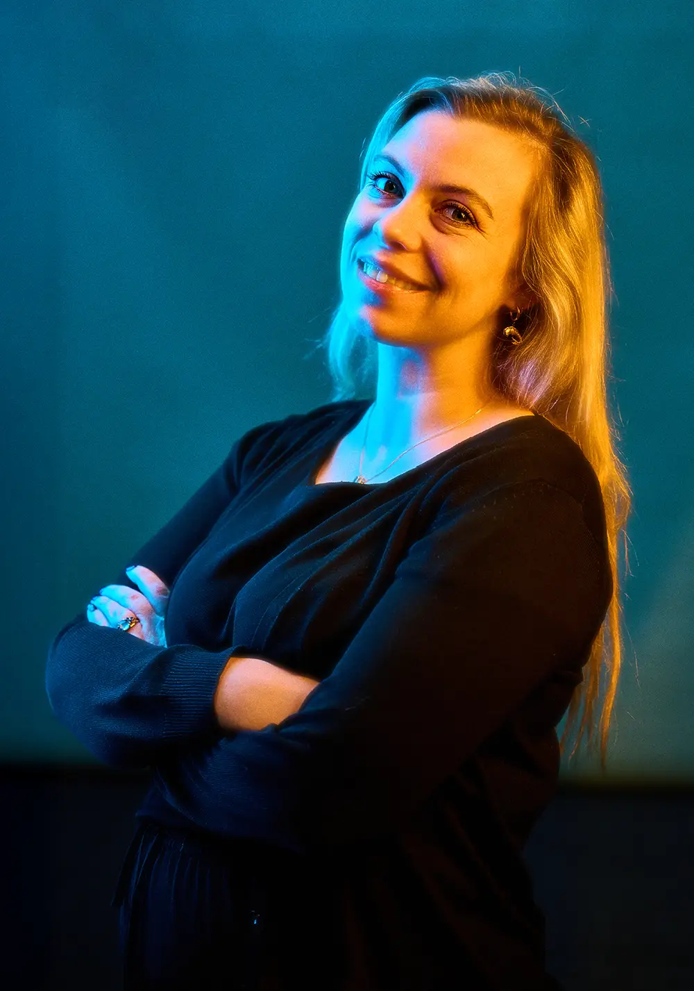 Smiling woman with long blonde hair wearing a black top, arms crossed, lit with blue and orange lights against a dark background.