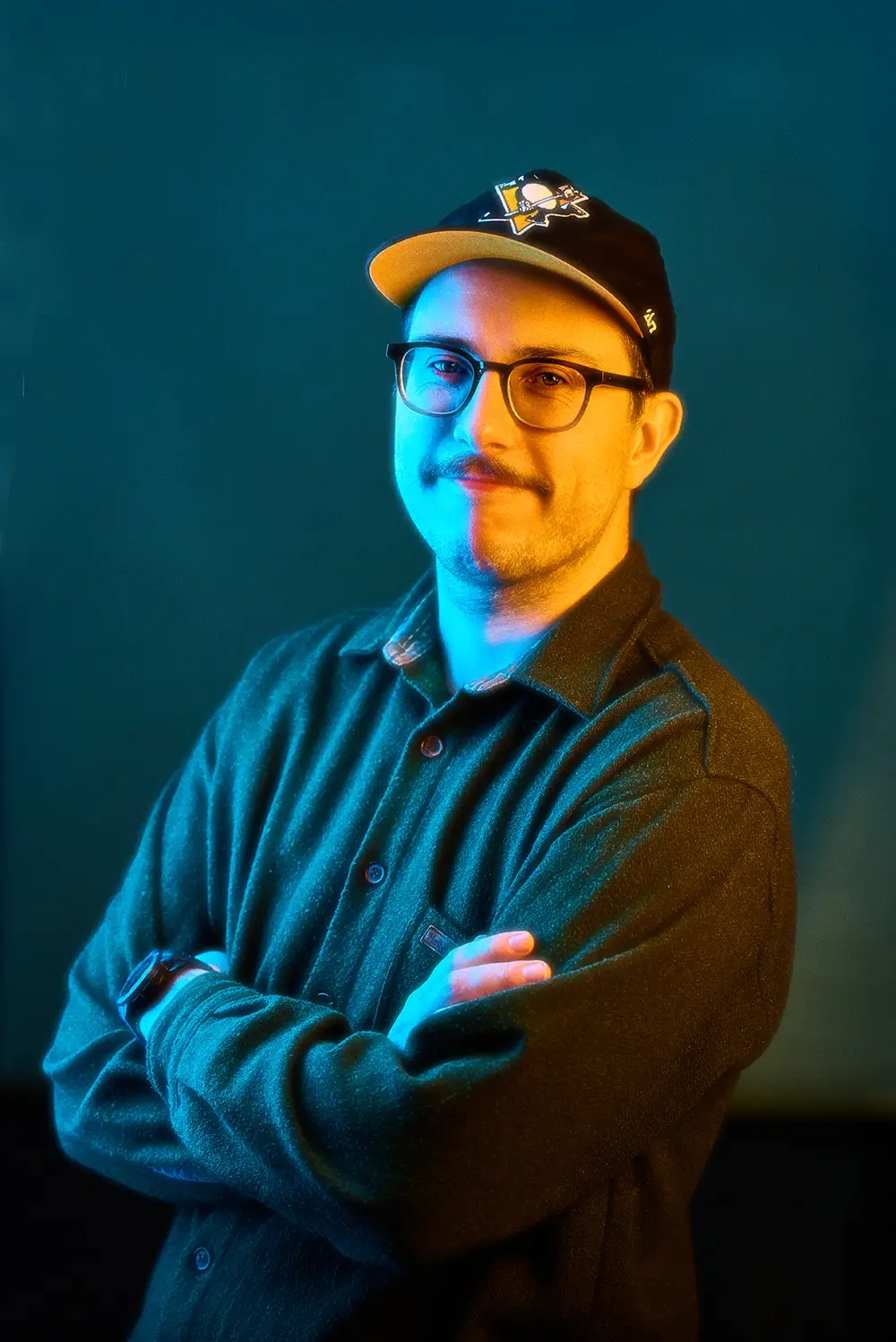 Man with glasses and mustache wearing a black cap and dark shirt, arms crossed, with blue and orange lighting.