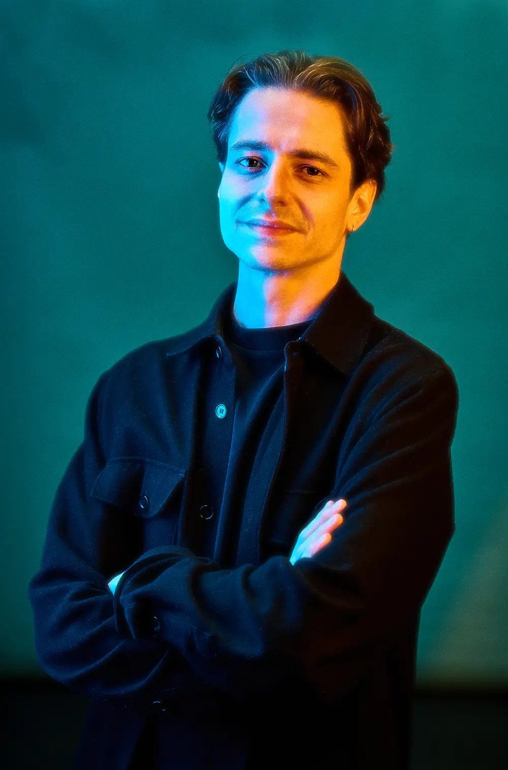 Man with short dark hair wearing a black jacket and crossing his arms, lit with blue and warm lighting against a teal background.