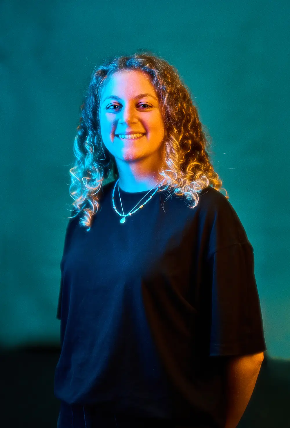 Smiling woman with curly blond hair wearing a black shirt and a necklace against a teal background.