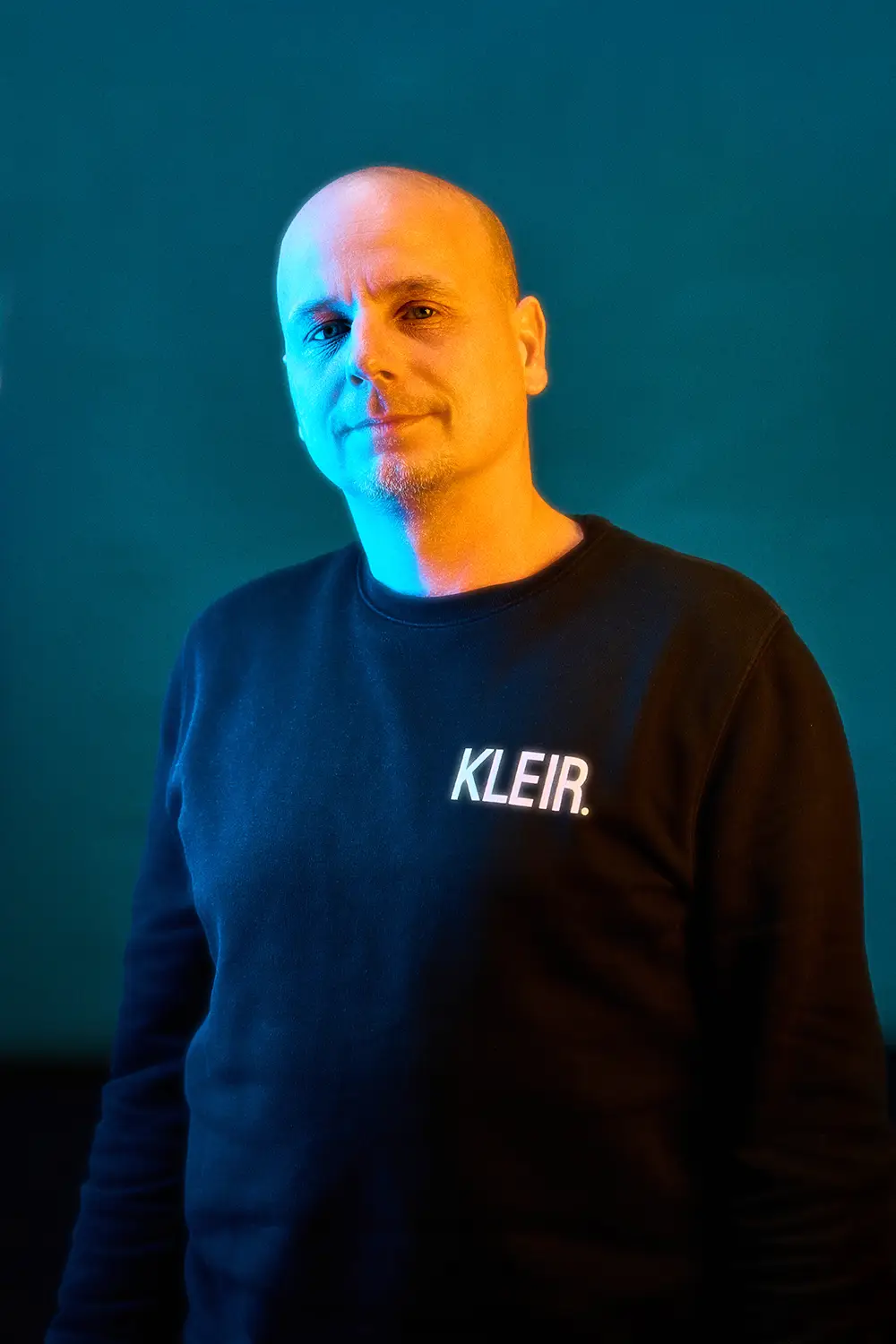 Man with a shaved head wearing a black sweater with the word KLEIR. illuminated by blue and orange lighting against a dark teal background.