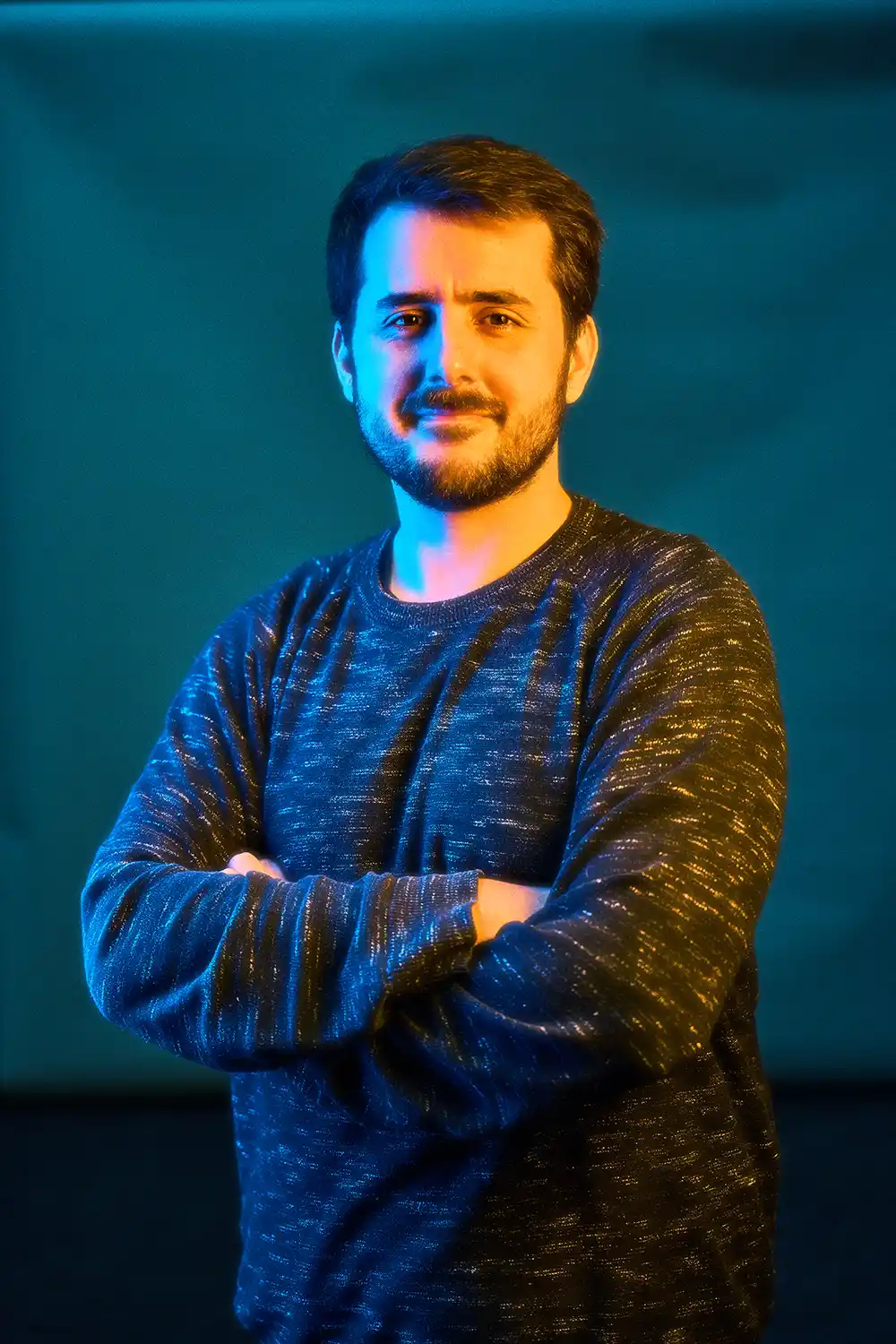 Man with beard and mustache standing with crossed arms, illuminated by blue and orange lighting on a dark background.
