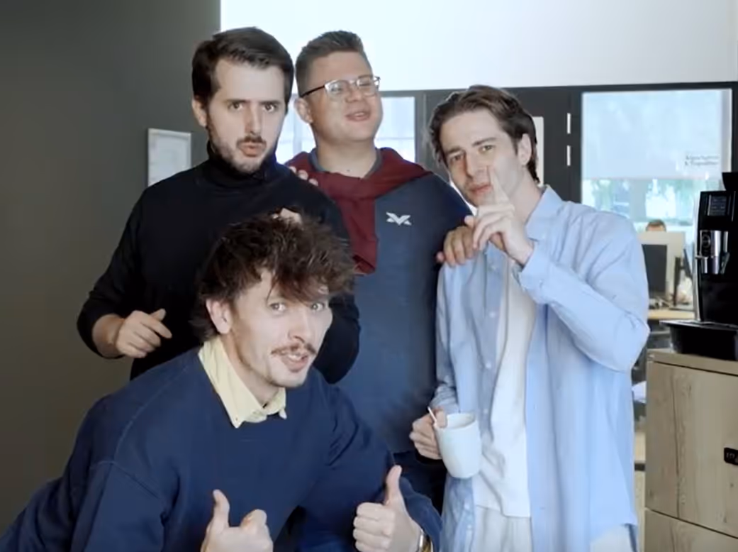 Four young men posing indoors, with one giving a thumbs-up and another holding a white mug.