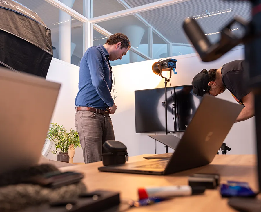 Man in a blue shirt standing with arms crossed, smiling in front of a camera in an office setting with another person working at a computer.