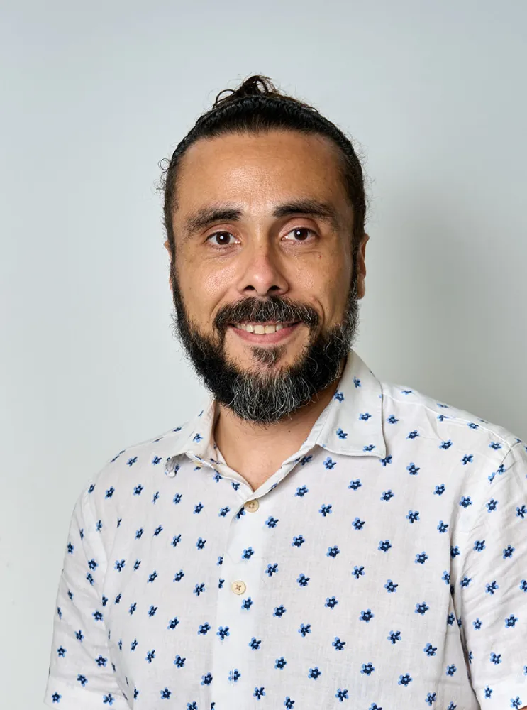 Hombre con barba y bigote, pelo recogido en un moño, vistiendo una camisa blanca con estampado de pequeñas flores azules, sonriendo frente a un fondo gris claro.