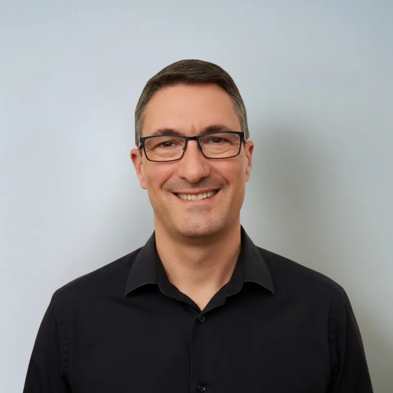 Hombre con gafas y camisa negra sonriendo frente a un fondo gris claro.