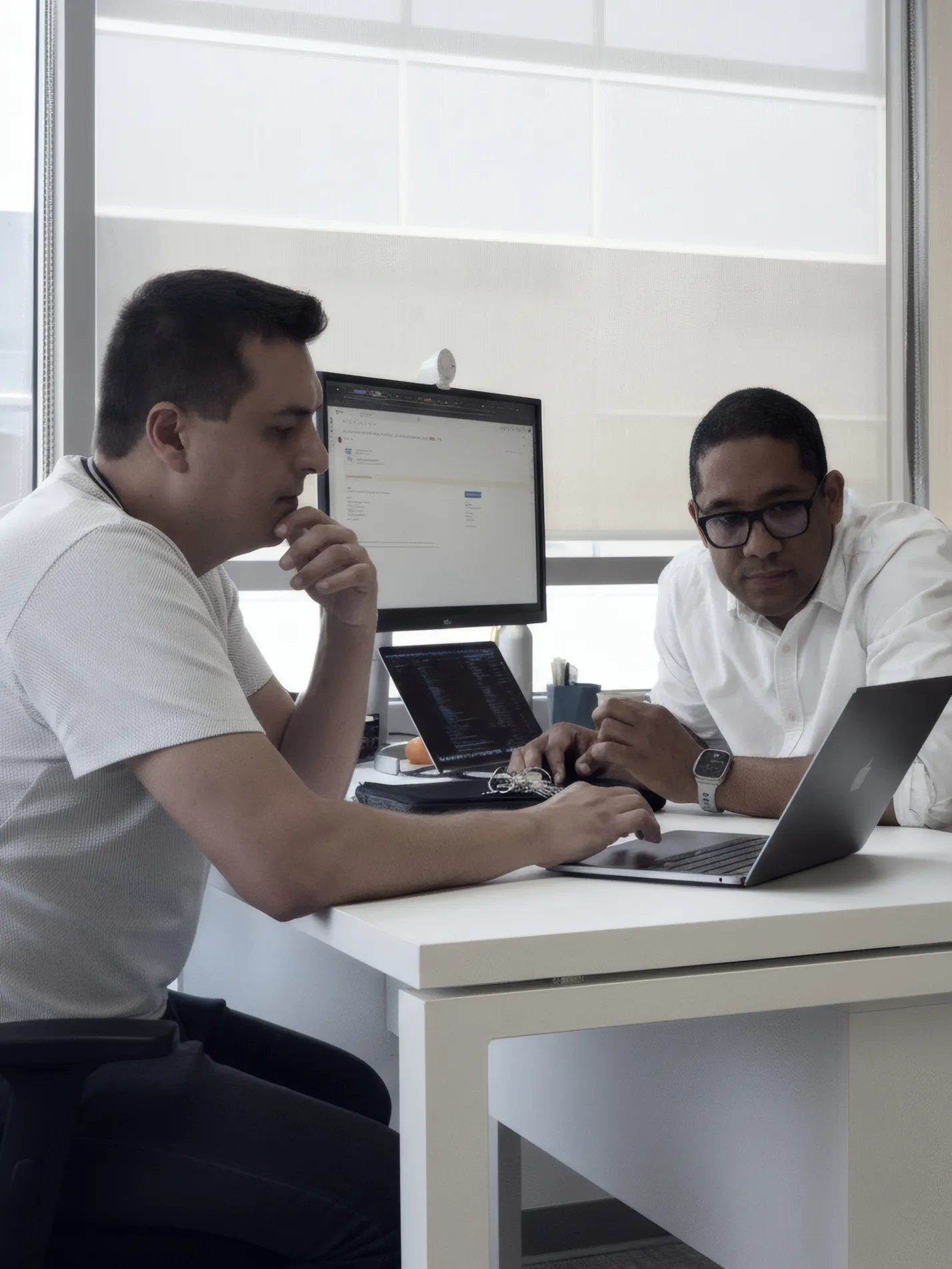 Dos hombres sentados juntos en una oficina mirando una computadora portátil sobre una mesa blanca mientras un monitor y una computadora portátil adicionales están al fondo.
