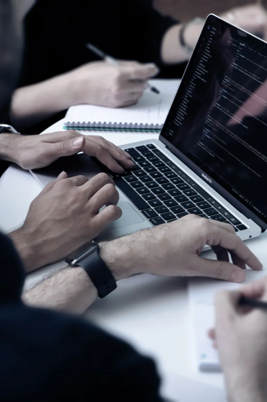 Manos de dos personas trabajando juntas frente a una computadora portátil con código en la pantalla, mientras una persona toma notas en un cuaderno.