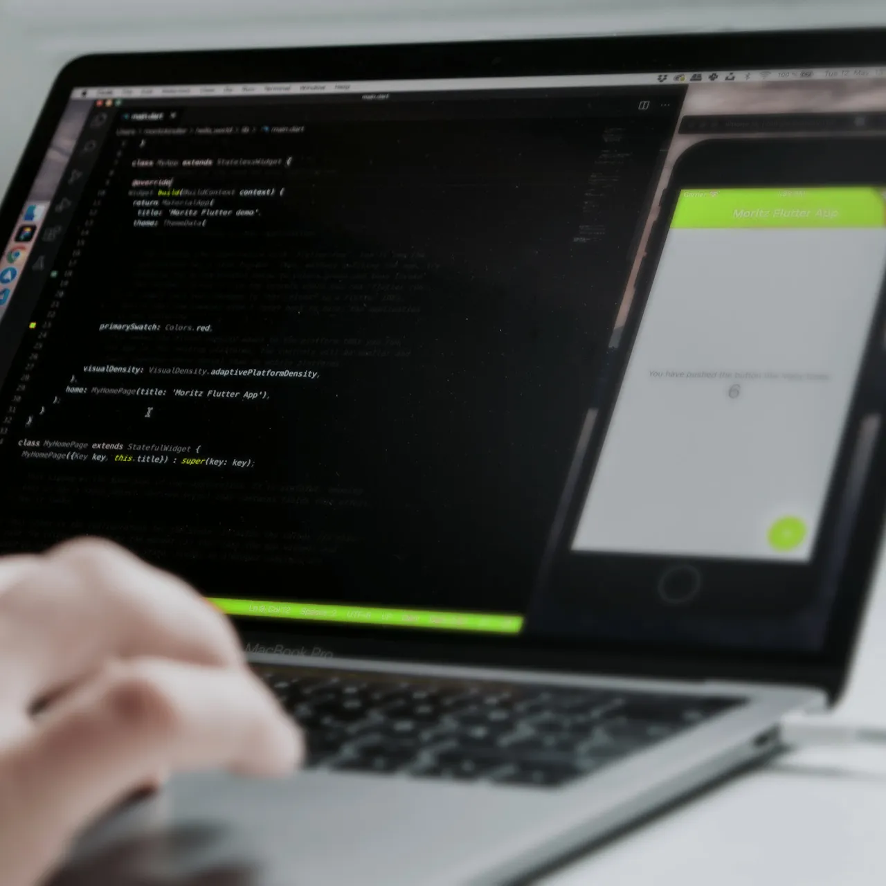 Mano sobre el teclado de una MacBook Pro con pantalla mostrando un IDE de programación y la vista simulada de una app Flutter de contador.