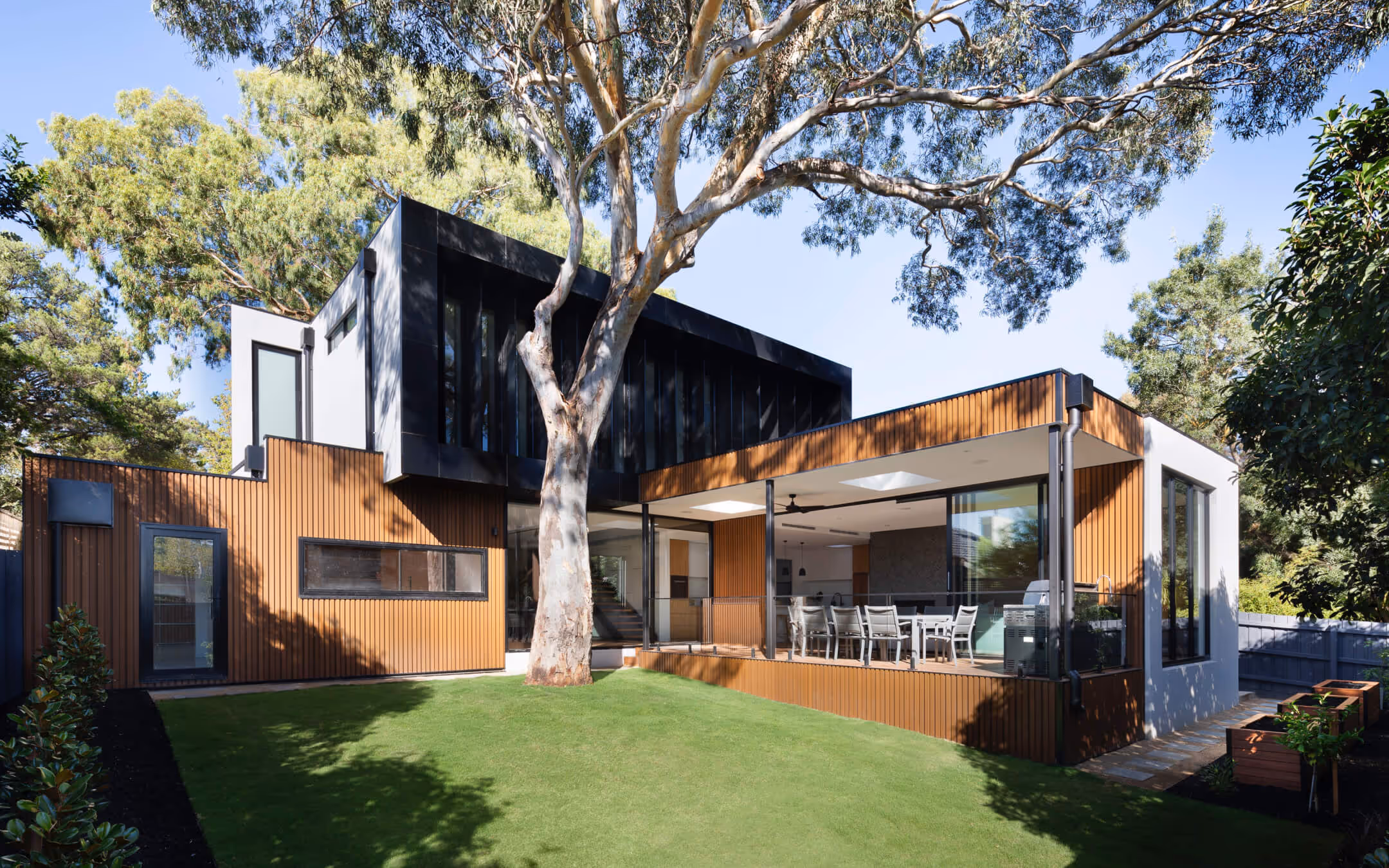 Maison moderne avec façade en bois et grandes fenêtres, entourée d'arbres et d'une pelouse verte bien entretenue.