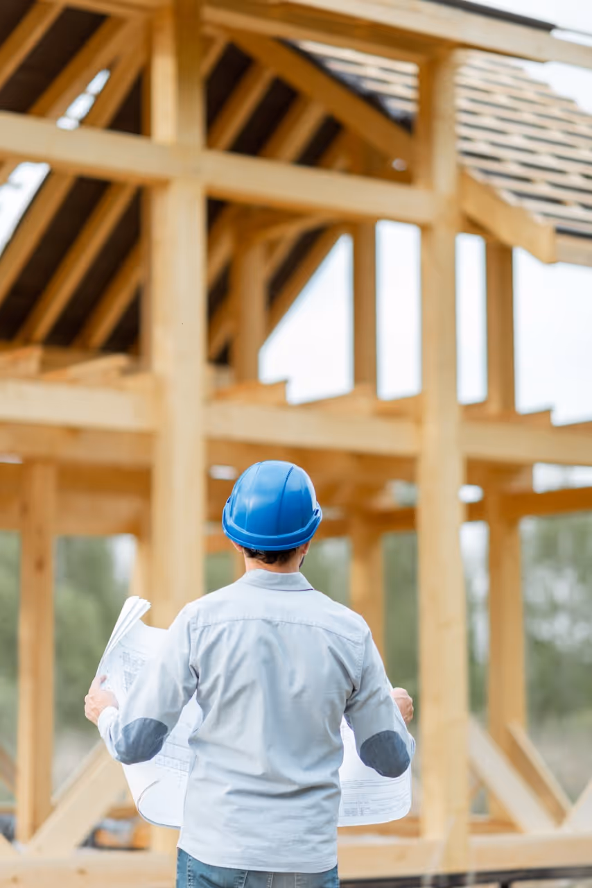 Homme portant un casque bleu regardant des plans devant une charpente en bois en construction.