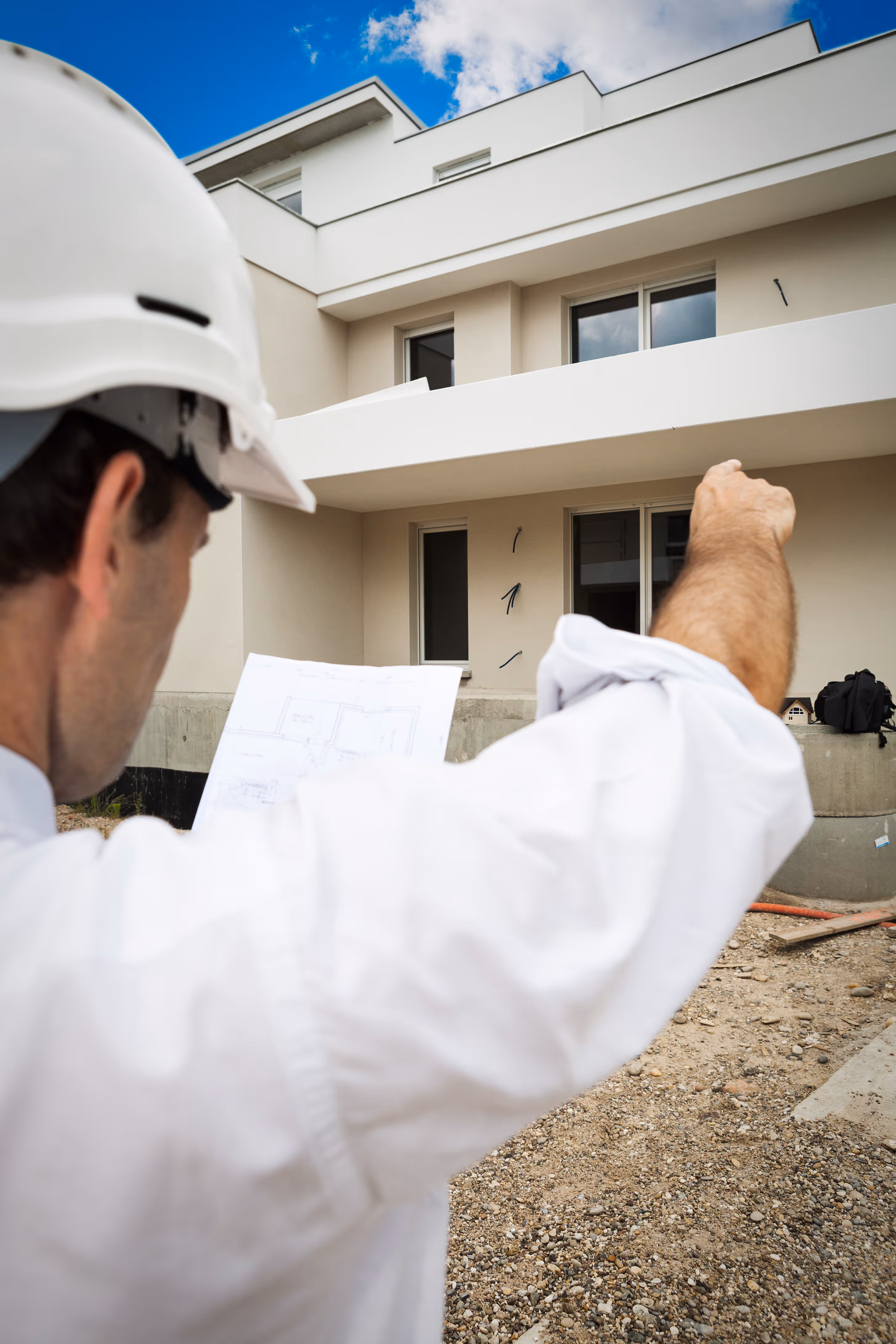 Un homme portant un casque de chantier tenant des plans et pointant vers un bâtiment résidentiel en construction.