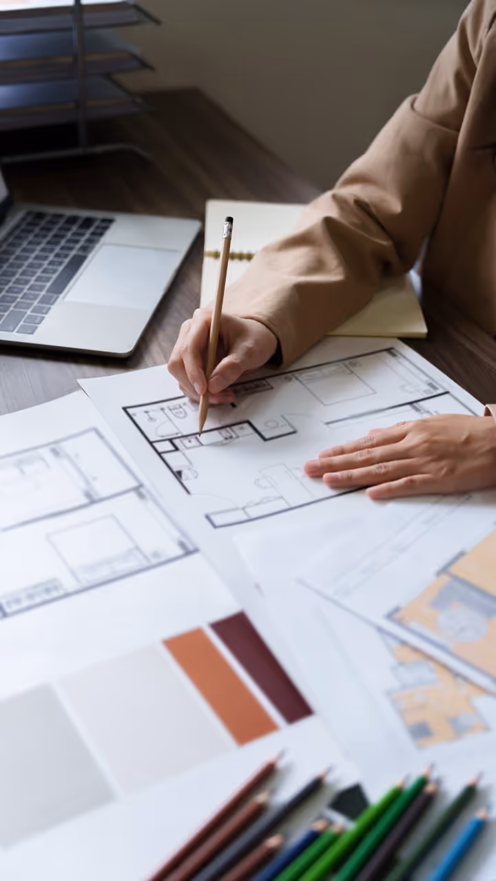 Person dessinant des plans d'architecture sur une table avec un crayon, entouré de plans de maison et d'échantillons de couleurs.