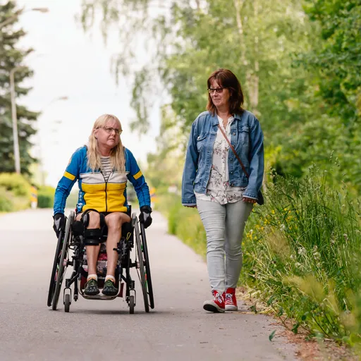 Två kvinnor är ute på promenad. En av dem sitter i rullstol.