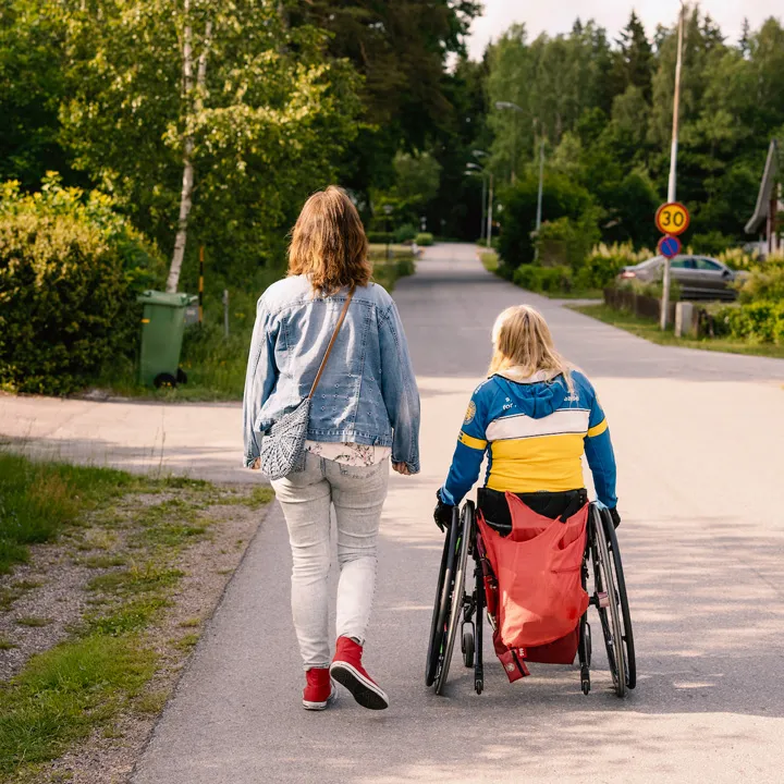 En person i rullstol och hennes assistent är ute och promenerar.