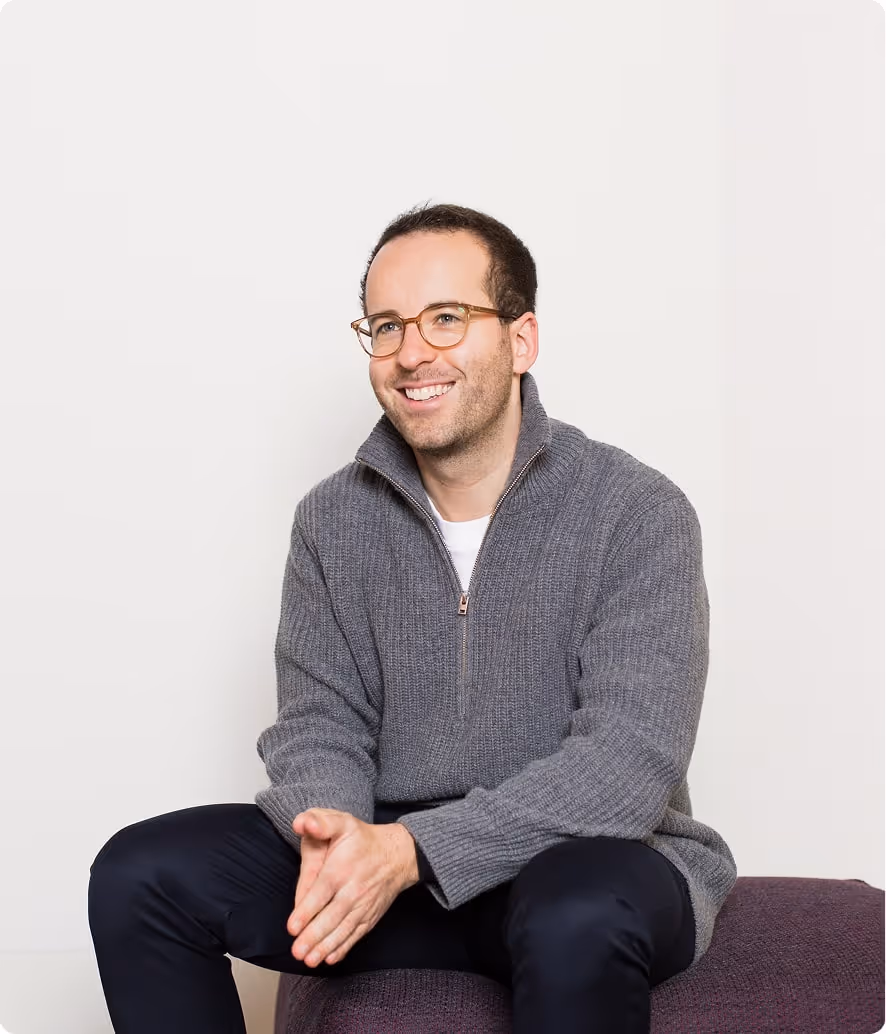 Smiling man wearing glasses and a gray sweater sitting on a purple ottoman against a plain white background.