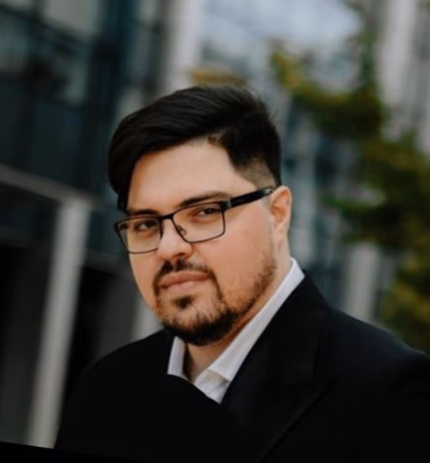 Man with dark hair, beard, and glasses wearing a black suit jacket and white shirt in an outdoor urban setting.