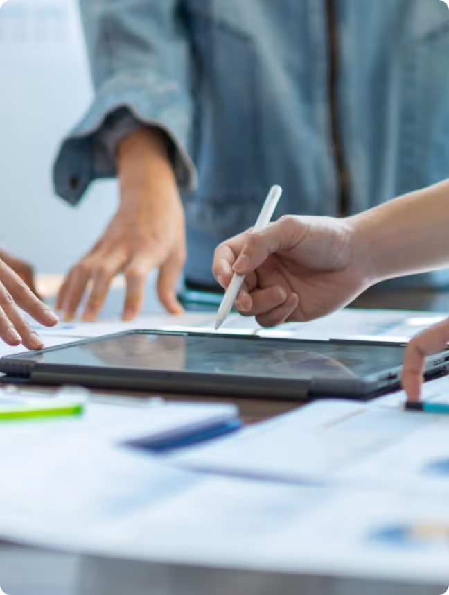 Close-up of hands collaborating over a tablet with a stylus and scattered documents on a table.