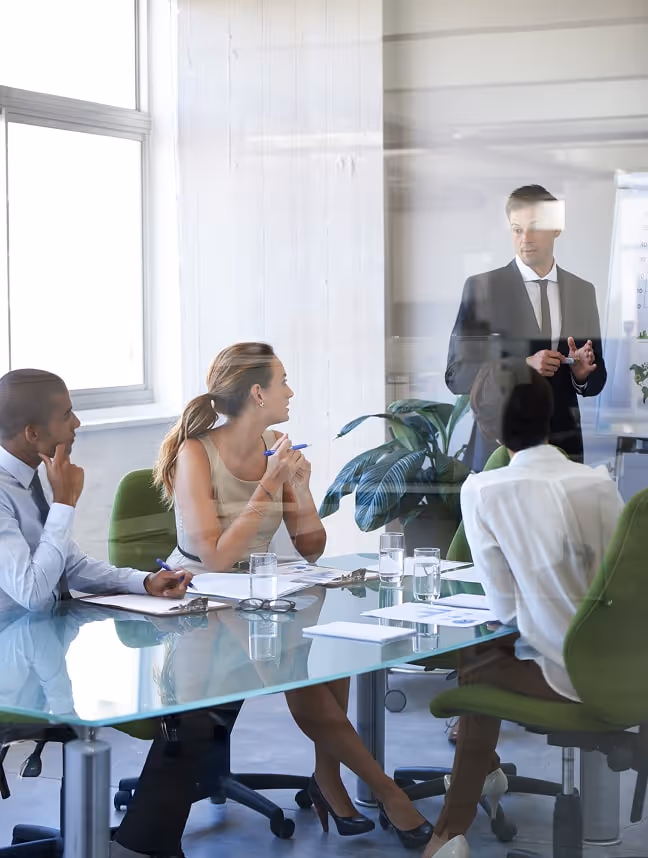 Businessman in suit presenting at a meeting table with three colleagues listening and taking notes in a bright office.