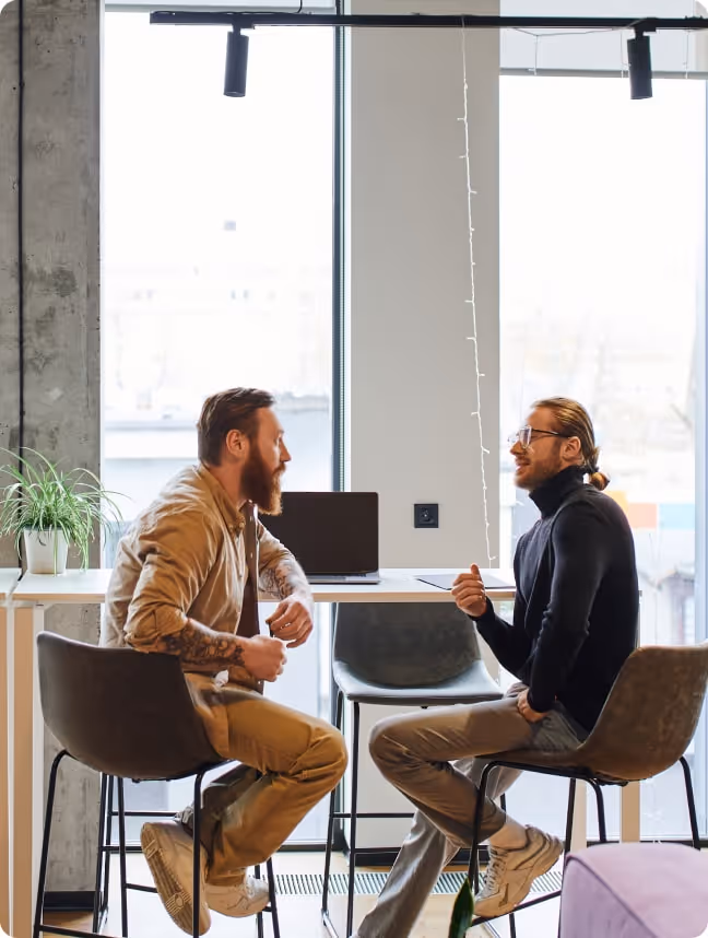 Two men engaged in conversation sitting on chairs by a window with a laptop and a plant on the table behind them.