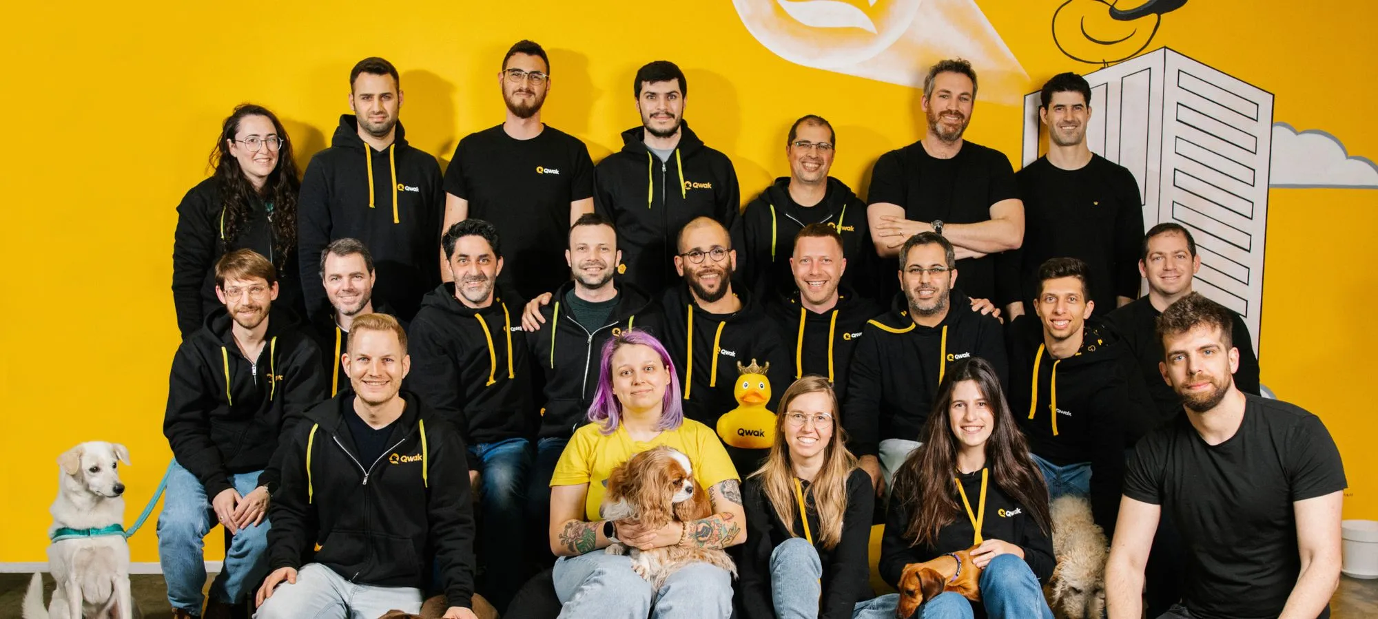 Group photo of twenty people wearing black Qwak hoodies against a yellow wall, with three dogs and one person holding a yellow rubber duck.