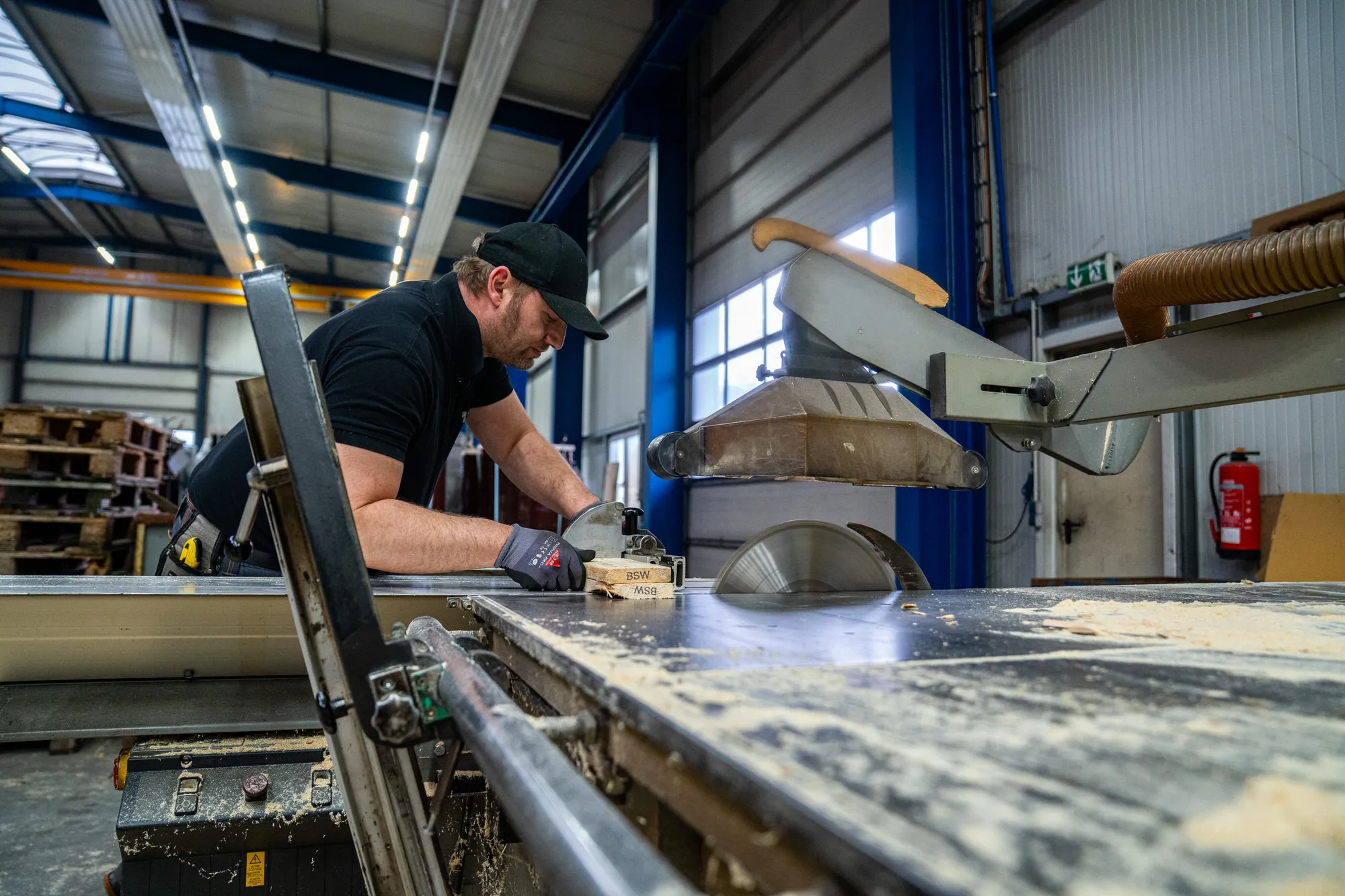 Man wearing a black T-shirt and cap operates a circular saw in a workshop with wooden pallets in the background.