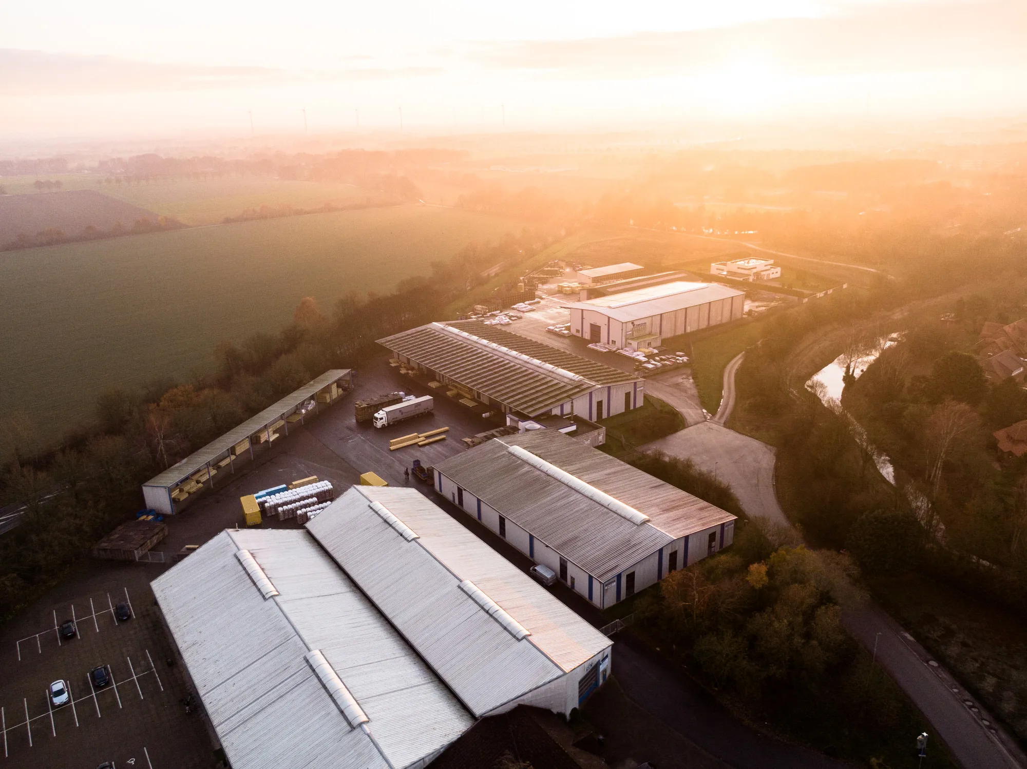 Aerial view of an industrial area with several warehouses, trucks and storage materials at sunrise with fields and wind turbines in the background.